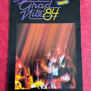 Può includere: Una fotografia in bianco e nero di una band che suona sul palco durante il Grad Nite '84 di Disneyland. La band suona davanti a uno sfondo luminoso e colorato. Il testo "Grad Nite '84" è in lettere viola e gialle. Il testo "KROQ" è in lettere gialle con sfondo rosso.