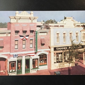 May include: A vintage postcard showing a street scene with two brick buildings. The building on the left has a red, white and blue flag hanging from the window. The building on the right has a sign that reads "Urjohn Pharmacy".