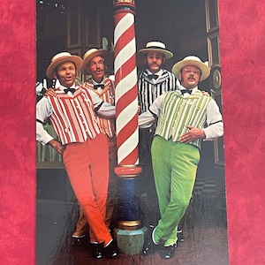 May include: Four men wearing striped vests and hats stand in front of a red and white striped barber pole. The men are smiling and looking at the camera. The barber pole is a classic symbol of the barbershop.