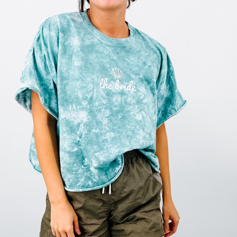 May include: A teal blue tie-dye t-shirt with a white seashell and the text "the bride" embroidered on the front.
