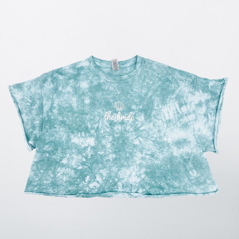 May include: A light blue tie-dye cropped t-shirt with a white seashell and the text "the bride" embroidered on the front.