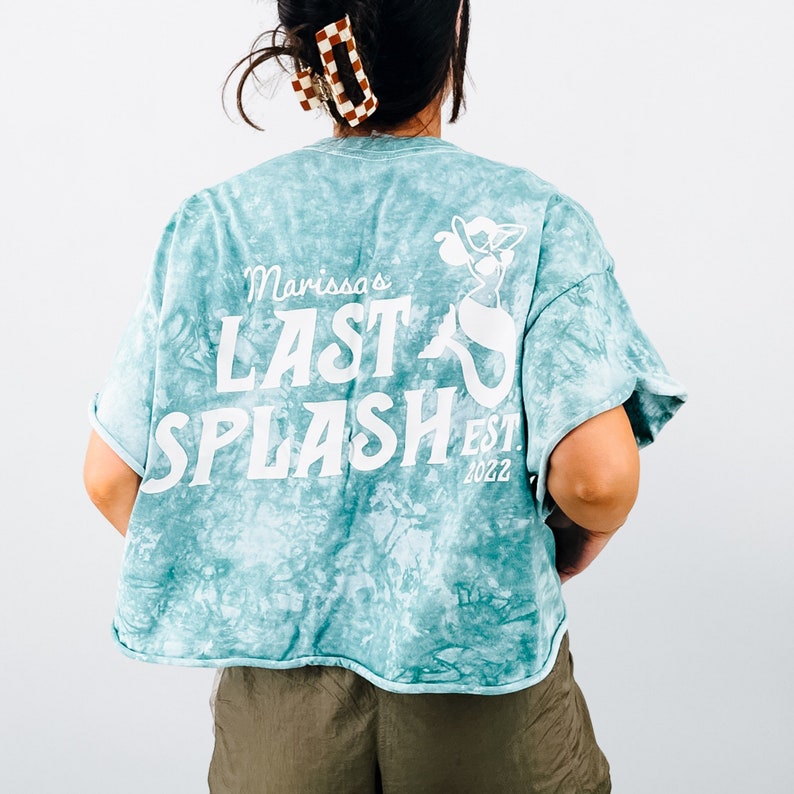 May include: A teal blue tie-dye cropped t-shirt with a white graphic of a mermaid and the text "Marissa's Last Splash Est. 2022".