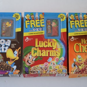 May include: Three boxes of General Mills cereal with free mini Pez dispensers. The boxes are Cocoa Puffs, Lucky Charms, and Honey Nut Cheerios. Each box features a cartoon character and the cereal mascot.