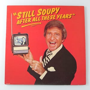 May include: Album cover with a red background featuring a man in a suit holding a small television. The text "Still Soupy After All These Years" is in yellow. The man is smiling and the television shows a black and white image.