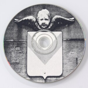 May include: A grayscale CD with a detailed graphic. The central image features a cherub with outstretched wings above a shield-like shape. The CD has text around the edges, including the record label and copyright information.