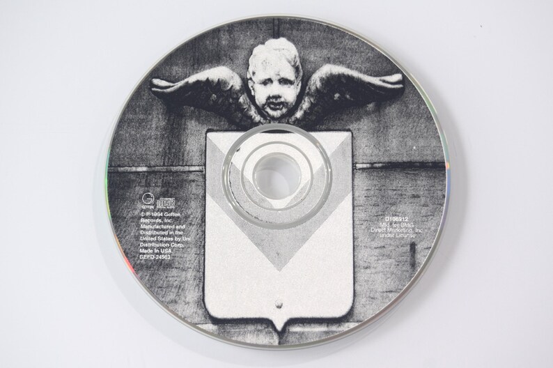 May include: A grayscale CD with a detailed graphic. The central image features a cherub with outstretched wings above a shield-like shape. The CD has text around the edges, including the record label and copyright information.