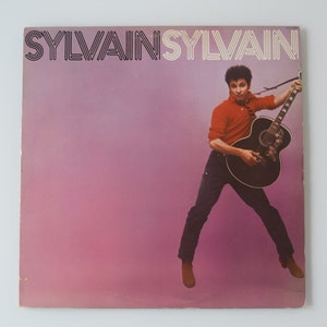 May include: A vintage vinyl record album cover featuring a man in a red shirt and blue jeans playing a black acoustic guitar. The album cover is pink and has the text "SYLVAIN SYLVAIN" in black.