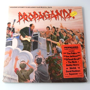 May include: Album cover for "Propaganda" featuring a band performing live. The artwork depicts a crowd of people with their hands raised, and the band is playing on a stage. The album cover has a red and orange color scheme.