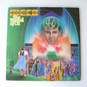 May include: A vintage vinyl record album cover for MECO's "The Wizard of Oz." The cover features a vibrant illustration with a large portrait of a man with a glowing head, surrounded by characters from the classic film, in a colourful, retro style.