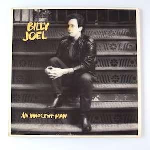 May include: Album cover for Billy Joel's "An Innocent Man." The cover features a black and white photo of Billy Joel sitting on a staircase. The album title and artist's name are in yellow text.