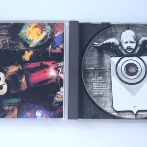 May include: Opened CD case with a disc featuring a grayscale image of a cherub with wings above a shield-like shape. The case's cover art displays a collage with the number "3" and colorful abstract elements.