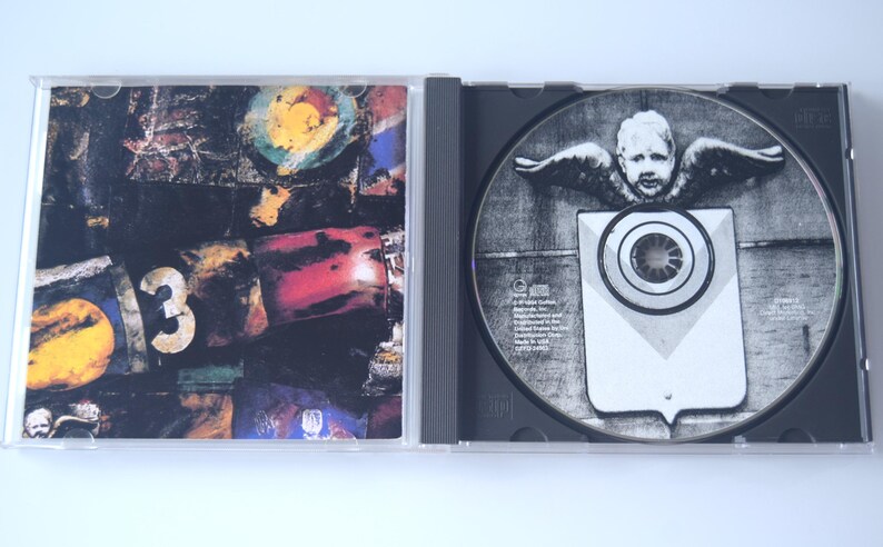 May include: Opened CD case with a disc featuring a grayscale image of a cherub with wings above a shield-like shape. The case's cover art displays a collage with the number "3" and colorful abstract elements.