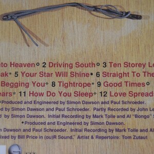 May include: A wooden surface with song titles and credits in red text. The titles include "Breaking Into Heaven," "Driving South," and "Ten Storey Love Song." A gray cord is at the top.