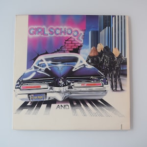 May include: A vintage vinyl record album cover featuring a black car with the text "Girl School" in pink neon letters and the text "Hit and Run" in white letters. Three women in black leather jackets are standing behind the car.