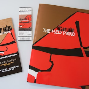 May include: An image featuring promotional materials for Elton John's "The Red Piano" show. The items include a program, a ticket, and a larger book, all with a red piano graphic and the show's title. The color scheme is red, brown, black, and white.