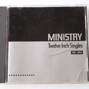 May include: A silver CD case with a black spine. The front of the case has the text "MINISTRY" in large black letters, followed by "Twelve Inch Singles" in smaller black letters. The bottom of the case has the text "1981-1984" in smaller black letters.