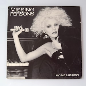 May include: A black and white photo of a woman with blonde hair holding a microphone. The photo is on a record album cover with the title "MISSING PERSONS" and the album title "RHYME & REASON".