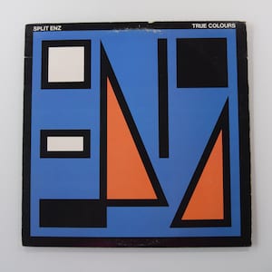 Peut inclure: Pochette d'album pour "True Colours" de Split Enz avec un design géométrique. La couverture a un fond bleu avec des formes noires, blanches et orange. Le nom du groupe et le titre de l'album sont imprimés en haut.