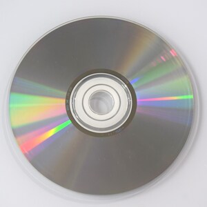 May include: A silver CD or DVD disc with a clear plastic case. The disc reflects a rainbow of colors, including green, yellow, and red. The center of the disc has a silver hub with a hole.