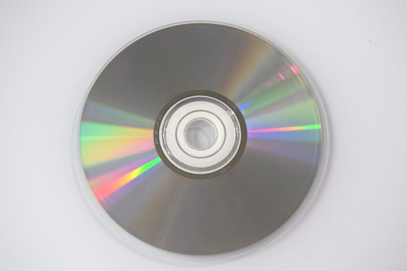 May include: A silver CD or DVD disc with a clear plastic case. The disc reflects a rainbow of colors, including green, yellow, and red. The center of the disc has a silver hub with a hole.