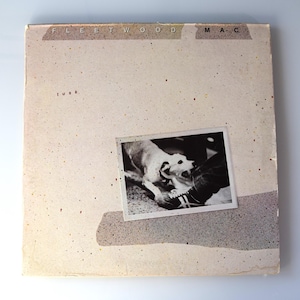 May include: Album cover for Fleetwood Mac's "Tusk." The cover is a beige color with the band's name at the top and the album title "tusk" below. A black and white photo of a dog biting a person's leg is in the center.