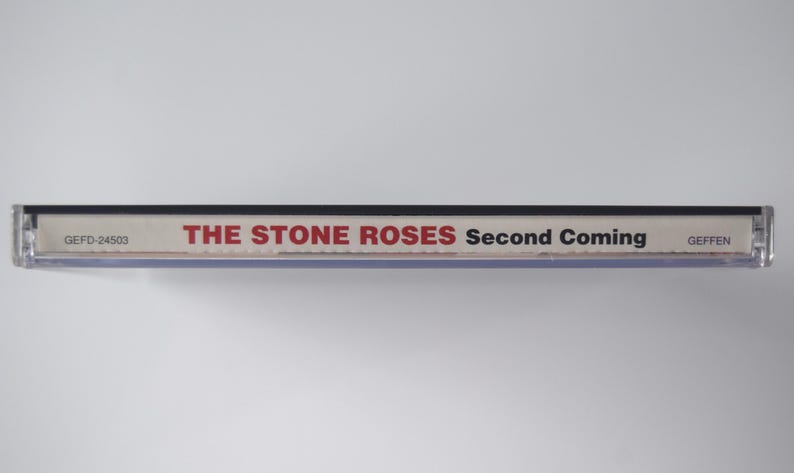 May include: A clear plastic CD case with the album title "Second Coming" by The Stone Roses. The text is in red and black. The case has the Geffen Records logo.