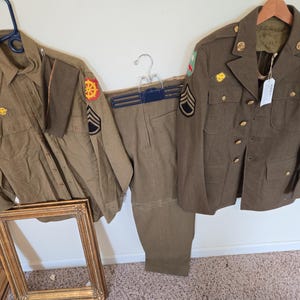 Pode incluir: Um conjunto de uniforme militar verde oliva, incluindo uma camisa abotoada, calças e um casaco. O casaco e a camisa apresentam patches e insígnias. O uniforme está pendurado em cabides. Uma moldura dourada está em primeiro plano.