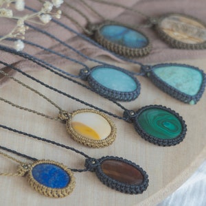 Può includere: Una collezione di otto collane con ciondoli in pietre preziose di diversi colori. Ogni ciondolo è avvolto in cordoncino macramé e ha una forma e un colore unici. Le collane sono esposte su una superficie in legno.