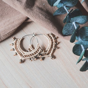 May include: A pair of gold and cream beaded hoop earrings with a silver hoop. The earrings are on a light wood surface with a brown fabric and green leaves in the background.