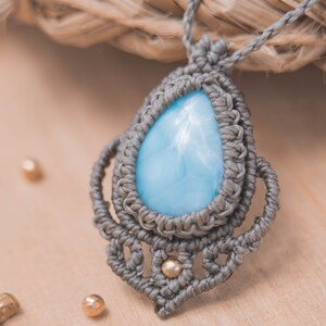 May include: A gray macrame necklace with a light blue teardrop-shaped gemstone pendant. The pendant is surrounded by intricate macrame knots and small gold beads.
