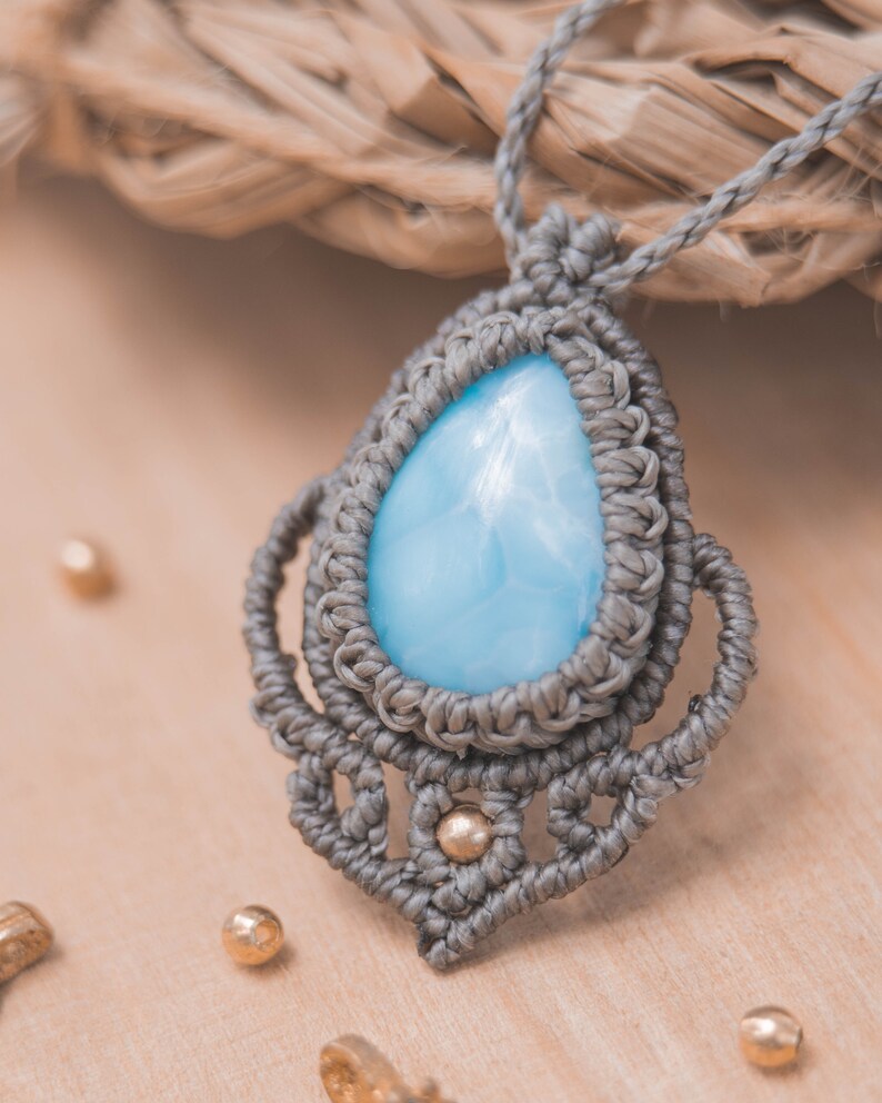 May include: A gray macrame necklace with a light blue teardrop-shaped gemstone pendant. The pendant is surrounded by intricate macrame knots and small gold beads.