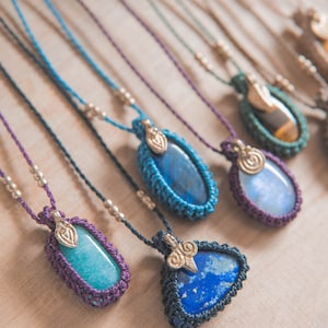 Puede incluir: Una colecci&oacute;n de collares de piedras preciosas hechos a mano. Cada collar presenta una piedra &uacute;nica en tonos azules, verdes y marrones, engastada en un marco de macram&eacute;. Los collares tienen cordones ajustables y detalles met&aacute;licos.