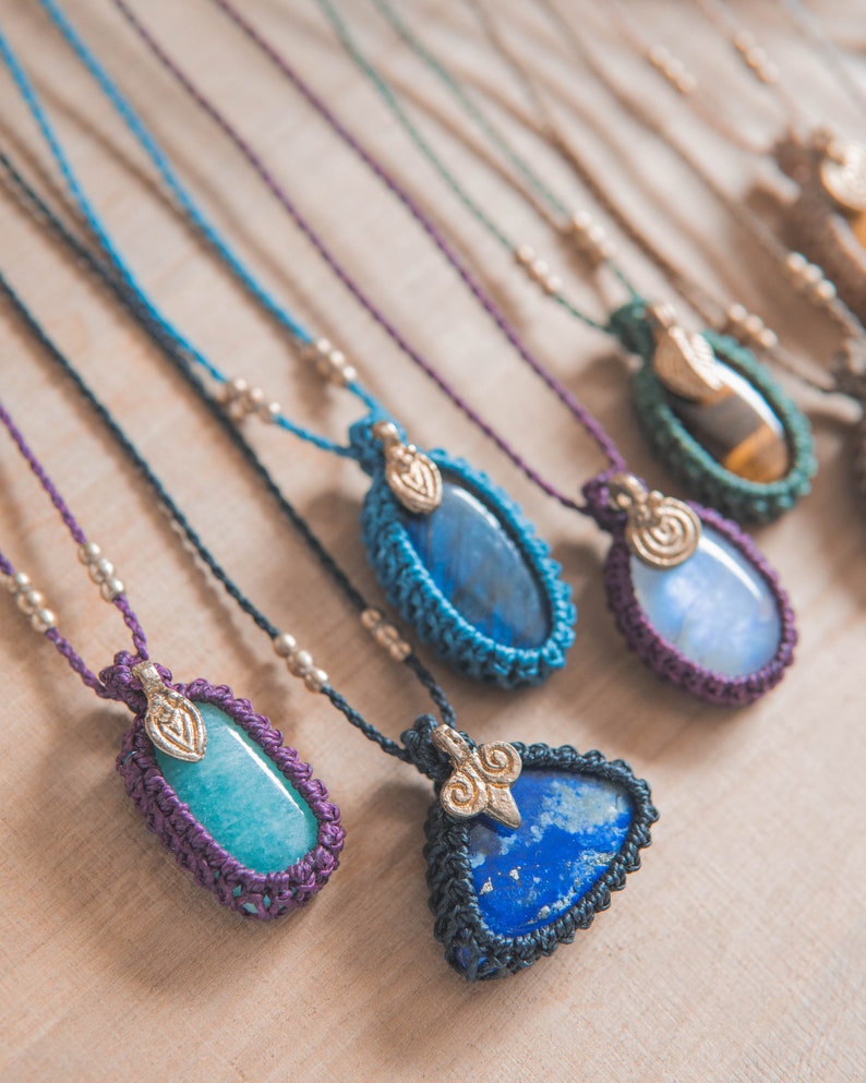 Puede incluir: Una colecci&oacute;n de collares de piedras preciosas hechos a mano. Cada collar presenta una piedra &uacute;nica en tonos azules, verdes y marrones, engastada en un marco de macram&eacute;. Los collares tienen cordones ajustables y detalles met&aacute;licos.