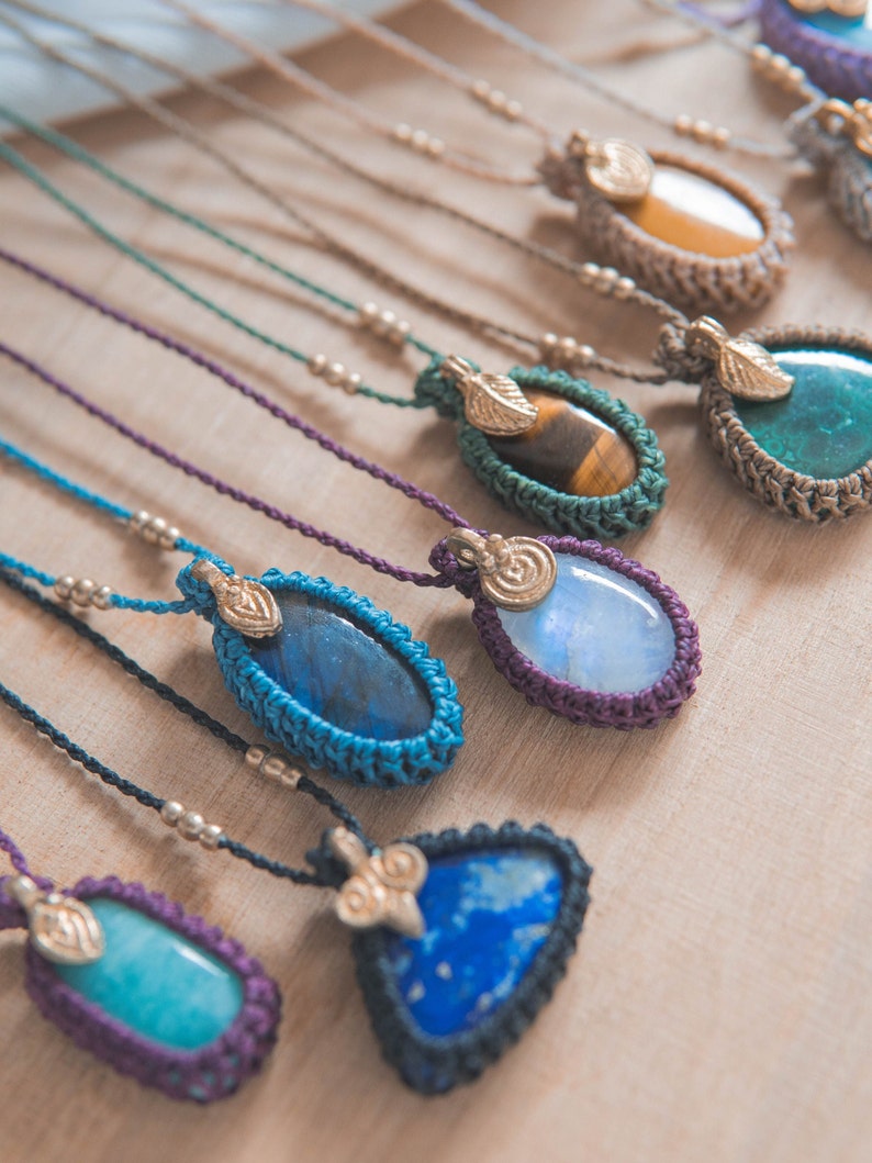 Puede incluir: Una colecci&oacute;n de collares con colgantes de piedras preciosas. Cada collar presenta una piedra &uacute;nica, incluyendo variedades azules, verdes y amarillas, engastada en un marco de ganchillo. Los colgantes cuelgan de un cord&oacute;n con peque&ntilde;as cuentas met&aacute;licas y una hoja dorada.