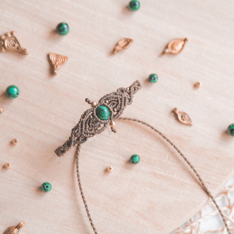May include: A green gemstone bracelet with a brown macrame design. The bracelet is on a wooden surface with small green and gold beads scattered around it.