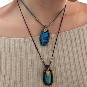 Puede incluir: Dos collares hechos a mano con colgantes de piedras preciosas. El collar superior tiene un colgante de piedra azul, mientras que el collar inferior presenta una piedra verde. Ambos colgantes est&aacute;n encapsulados en macram&eacute; y adornados con cuentas doradas.