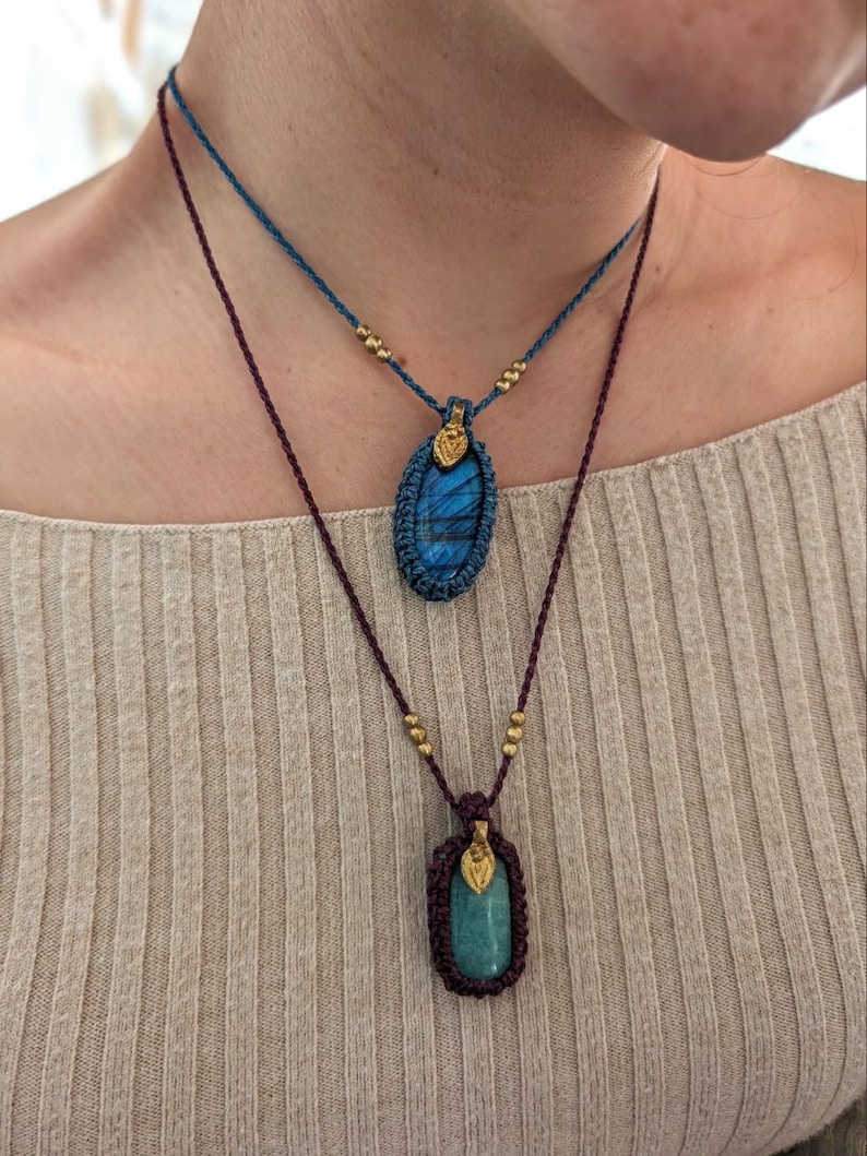 Puede incluir: Dos collares hechos a mano con colgantes de piedras preciosas. El collar superior tiene un colgante de piedra azul, mientras que el collar inferior presenta una piedra verde. Ambos colgantes est&aacute;n encapsulados en macram&eacute; y adornados con cuentas doradas.