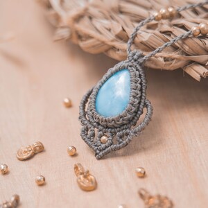 May include: A light blue gemstone pendant necklace with a gray macrame design. The necklace is on a wooden surface with gold beads scattered around it.