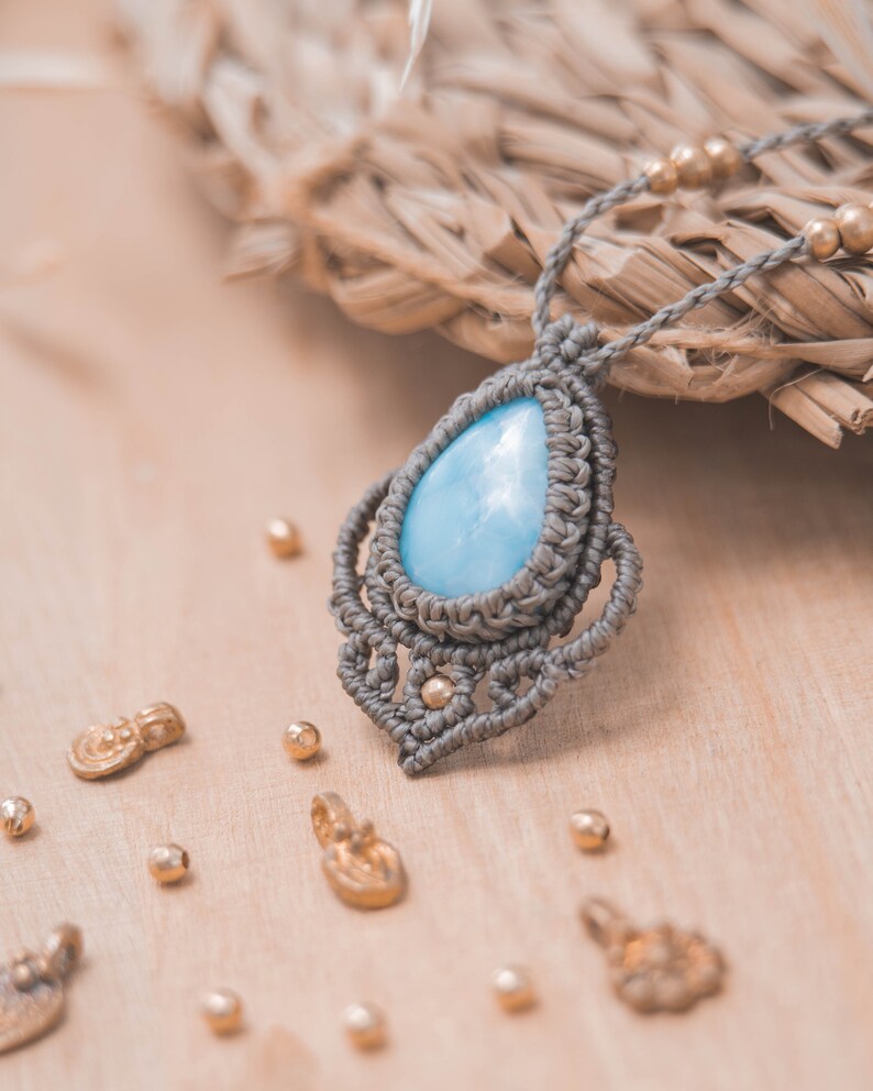 May include: A light blue gemstone pendant necklace with a gray macrame design. The necklace is on a wooden surface with gold beads scattered around it.