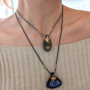 Puede incluir: Dos collares con colgantes de piedras preciosas. El collar superior tiene un cord&oacute;n verde y un colgante ovalado con una piedra ojo de tigre. El collar inferior tiene un cord&oacute;n negro y un colgante triangular de piedra azul. Ambos tienen detalles dorados.