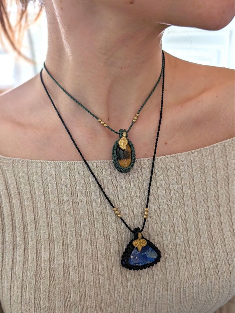 Puede incluir: Dos collares con colgantes de piedras preciosas. El collar superior tiene un cord&oacute;n verde y un colgante ovalado con una piedra ojo de tigre. El collar inferior tiene un cord&oacute;n negro y un colgante triangular de piedra azul. Ambos tienen detalles dorados.