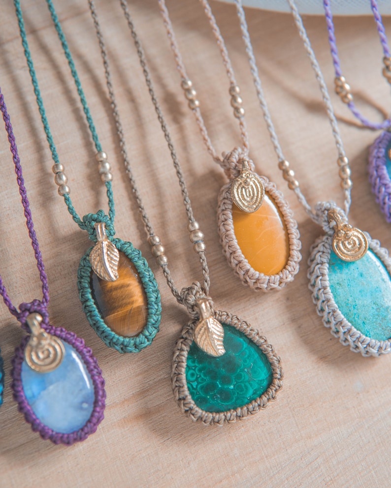 Puede incluir: Una colecci&oacute;n de collares de piedras preciosas hechos a mano. Cada collar presenta una piedra &uacute;nica de forma ovalada o de l&aacute;grima en varios colores, incluyendo azul, verde, amarillo y marr&oacute;n, engastada en un dise&ntilde;o de macram&eacute;. Los colgantes est&aacute;n adornados con dijes de hojas doradas y ensartados en collares de cord&oacute;n.