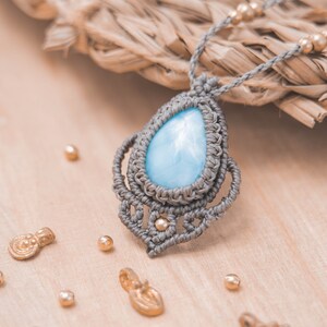 May include: A light blue gemstone pendant necklace with a gray macrame cord and gold accents. The pendant is in the shape of a teardrop and is surrounded by a delicate macrame design.