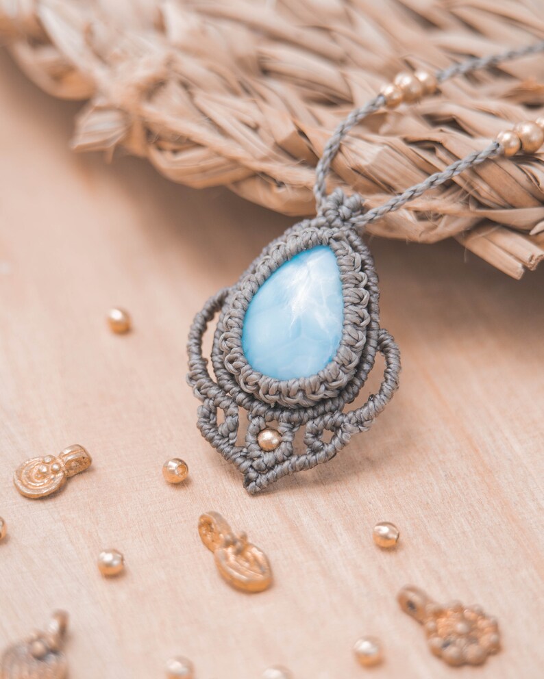 May include: A light blue gemstone pendant necklace with a gray macrame cord and gold accents. The pendant is in the shape of a teardrop and is surrounded by a delicate macrame design.