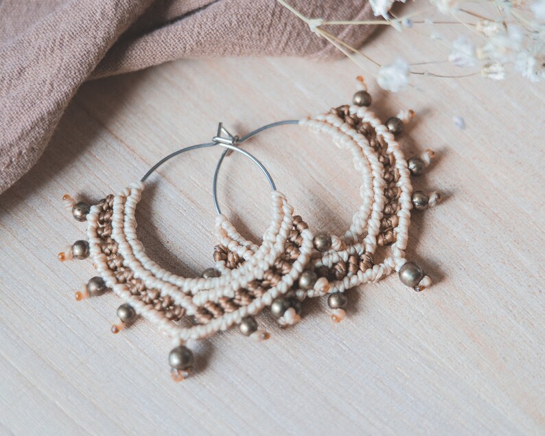 May include: A pair of hoop earrings with a macrame design in beige and brown with gold beads.