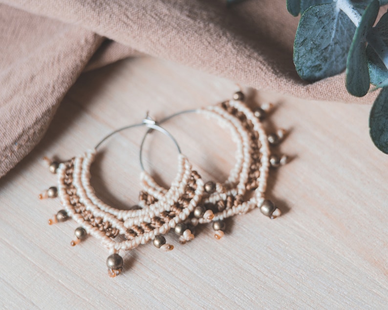 May include: A pair of hoop earrings with a woven design and gold beads. The earrings are made of a light brown and white material.