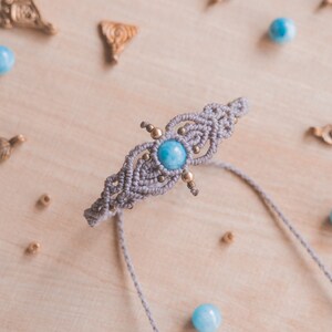 May include: A gray macrame bracelet with a light blue gemstone and gold beads. The bracelet is on a wooden surface with other beads and jewelry.