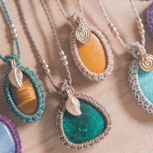 Puede incluir: Primer plano de varios collares de macram&eacute; con colgantes de piedras preciosas. Los colgantes son de varios colores, incluyendo verde, amarillo y marr&oacute;n. Cada collar tiene un dije de hoja y detalles de cuentas. Los collares se exhiben sobre una superficie de madera clara.