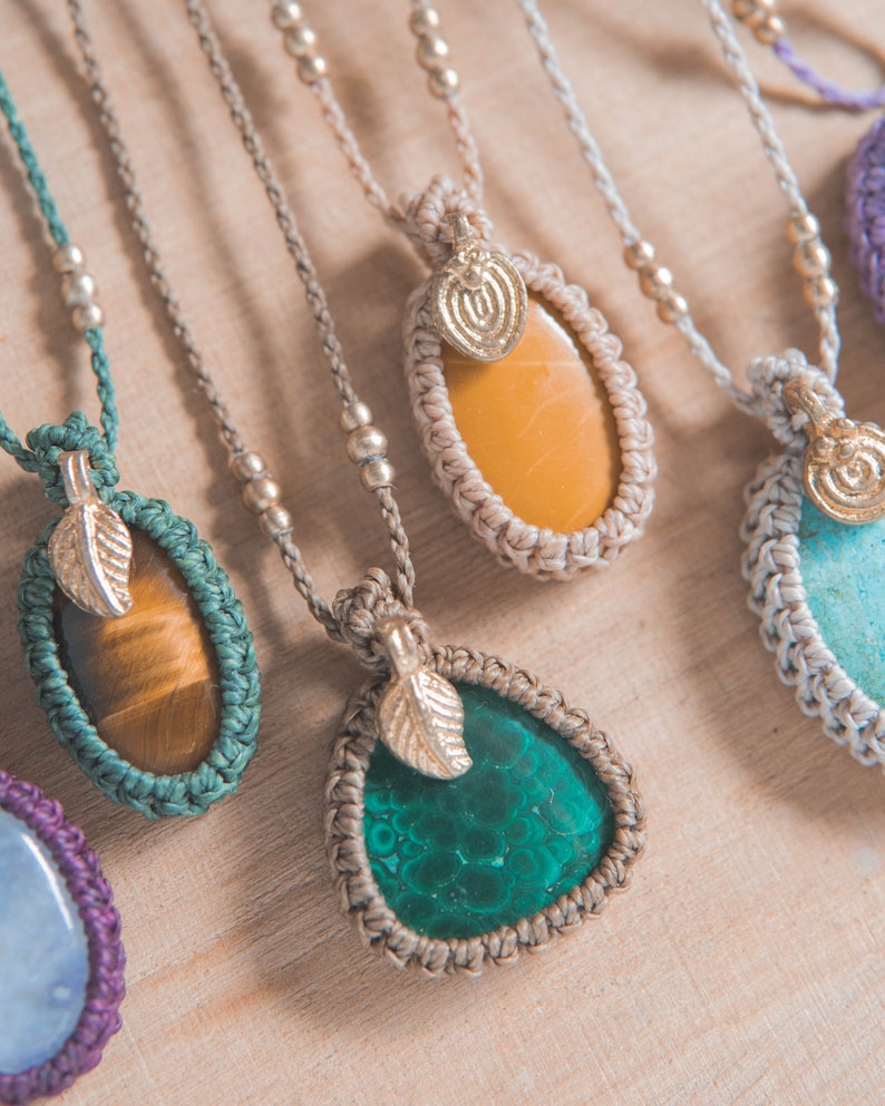 Puede incluir: Primer plano de varios collares de macram&eacute; con colgantes de piedras preciosas. Los colgantes son de varios colores, incluyendo verde, amarillo y marr&oacute;n. Cada collar tiene un dije de hoja y detalles de cuentas. Los collares se exhiben sobre una superficie de madera clara.