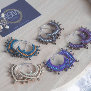 May include: Five pairs of handmade macrame hoop earrings with different colored threads and brass beads. The earrings are displayed on a wooden surface.